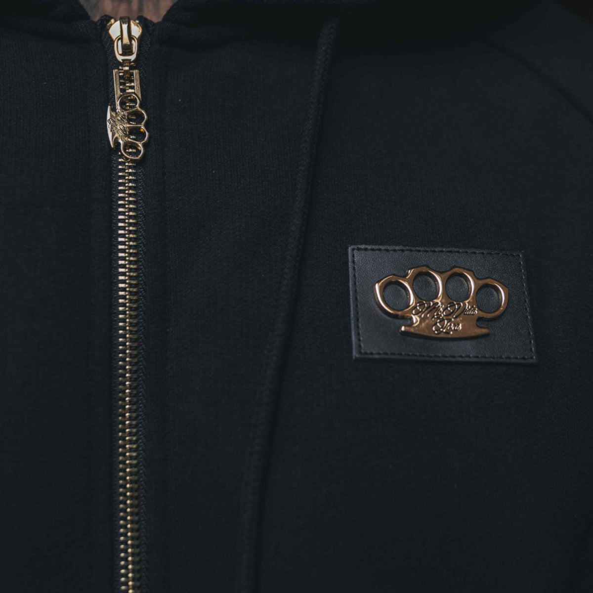 Nahaufnahme des goldenen MVL-Logos auf Lederpatch und des goldenen Reißverschlusses des Meander Hoodies