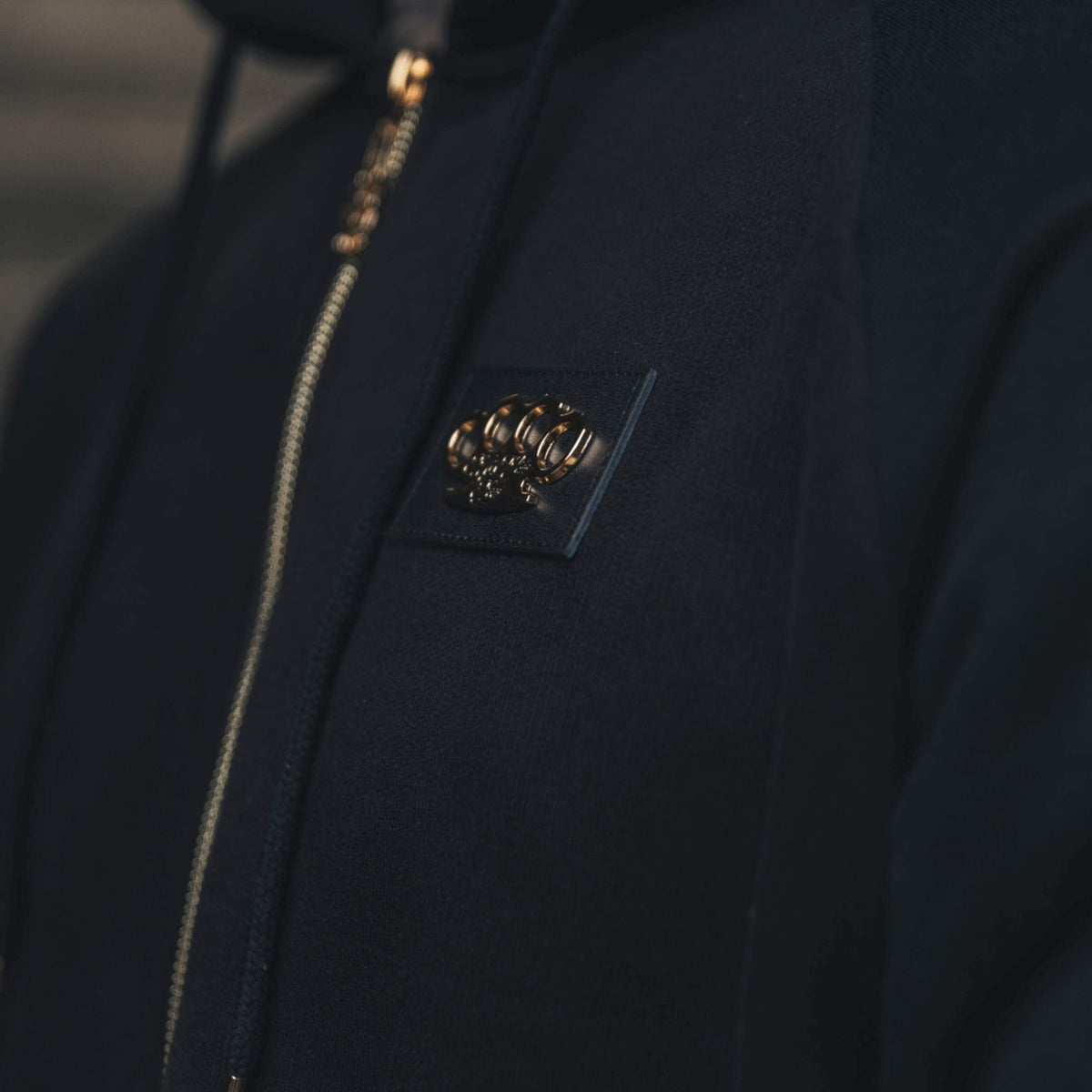 Nahaufnahme des goldenen MVL-Logos auf der Brust und des Reißverschlusses des schwarzen Meander Hoodies