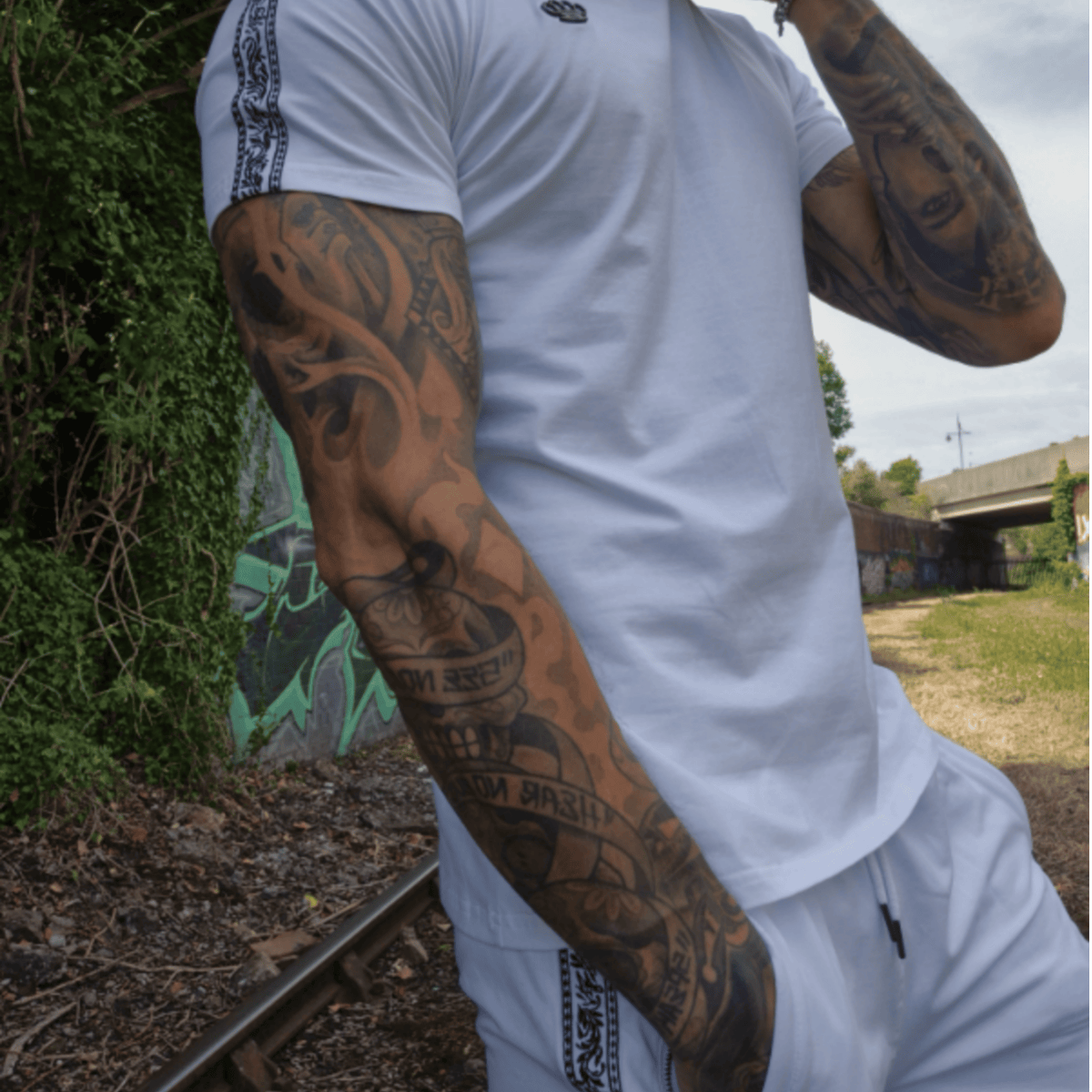 Model in wit MVL T-shirt en witte Meander korte broek bij spoorrails met graffiti