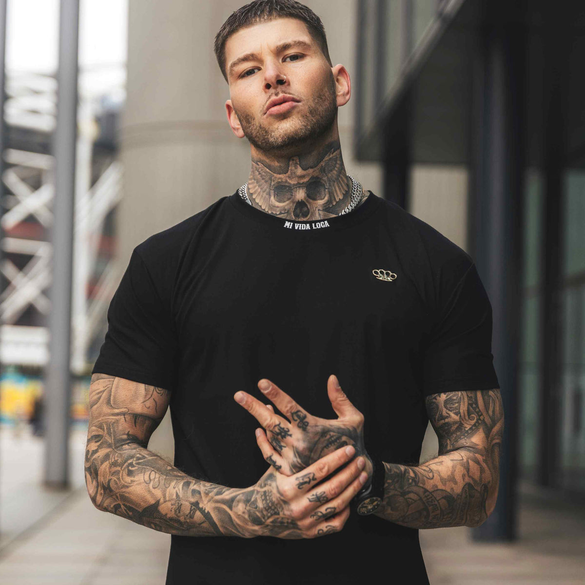 Tattooed model in the black FTS T-shirt, front, with hands folded in front of the chest