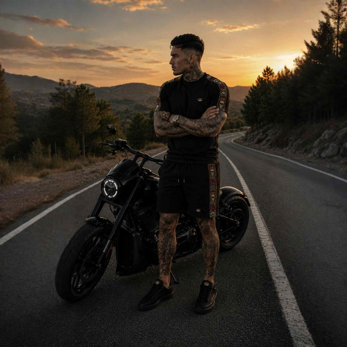 T-shirt Calavera nera indossata da modello a braccia conserte vicino a una moto nera su una strada di montagna