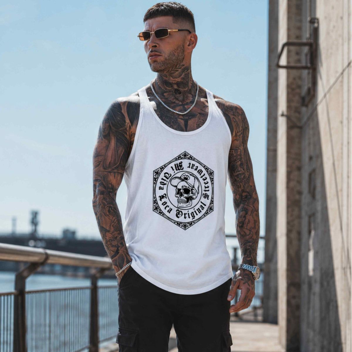 Modelo con tatuajes y gafas de sol lleva la camiseta de tirantes MVL Original streetwear blanca junto al agua