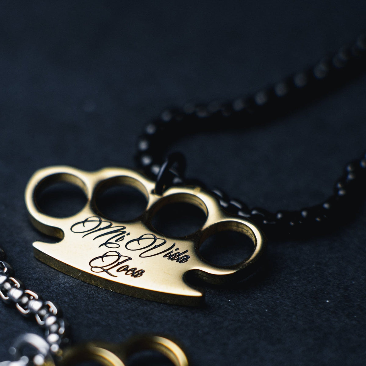 Close-up van de gouden MVL logo hanger met gegraveerde Mi Vida Loca tekst aan de zwarte ketting